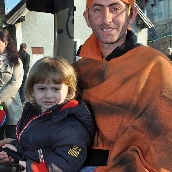Ein Mann in einem orangefarbenen Kostüm mit gelbem Stirnband posiert mit einem kleinen Mädchen in einer schwarzen Jacke. Sie sitzen auf einer Bank vor einem Gebäude.