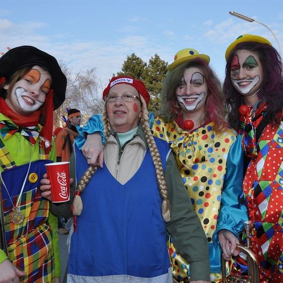Fünf Menschen in Clownkostümen stehen zusammen und einer hält einen roten Becher. Sie lächeln und scheinen für ein Foto zu posieren. Hinter ihnen ist ein verschwommener Hintergrund mit Bäumen und einer Straßenlaterne.