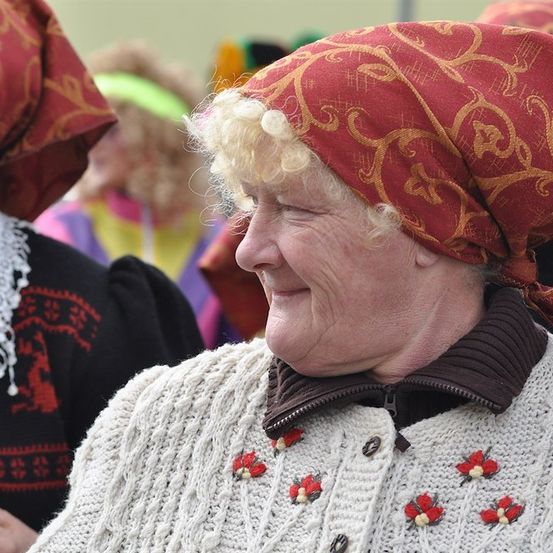Eine ältere Frau mit blonden Haaren trägt ein rot gemustertes Kopftuch und einen weißen Pullover mit roten Blumen und lächelt in die Kamera.