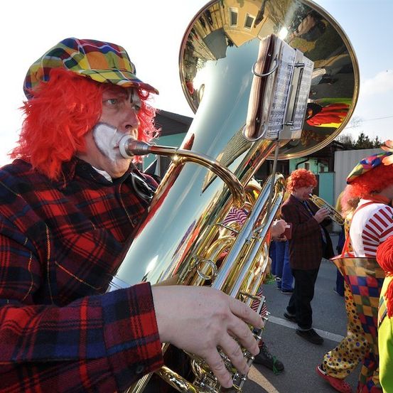 Ein Mann in Clown-Make-up spielt Tuba, während andere in Clown-Kostümen hinter ihm auf einem Außengelände stehen.