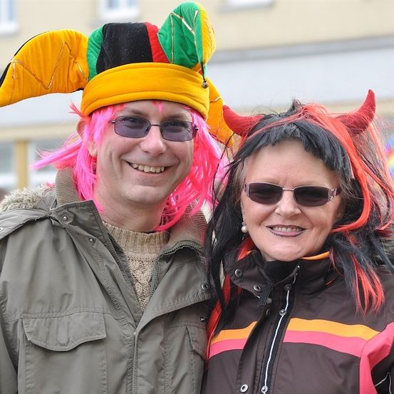Ein Mann und eine Frau in bunten Kostümen und Perücken, lächeln für die Kamera. Der Mann trägt eine Narrenmütze und eine rosa Perücke, während die Frau rote Teufelshörner und eine gestreifte Jacke hat.