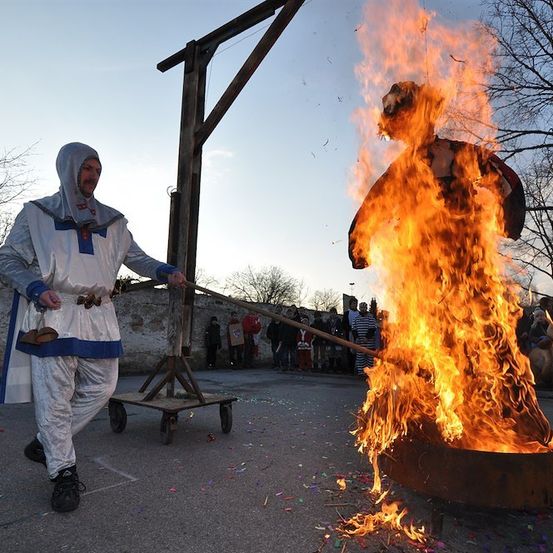 Ein Mann in einer weißen Robe steht neben einer brennenden Puppe in einem großen Metalltopf, während Zuschauer im Hintergrund zusehen.