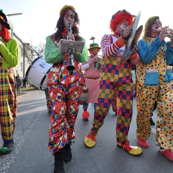Eine Gruppe von Clowns in bunten Kostümen und mit geschminkten Gesichtern spielt Musikinstrumente bei einer Parade.