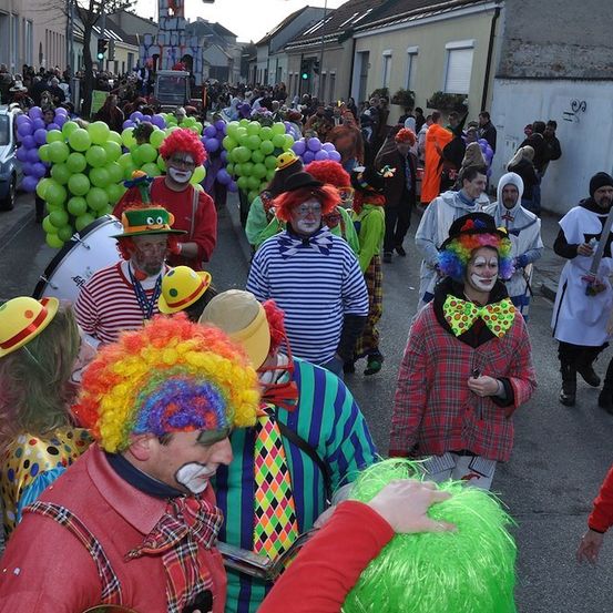 Eine Straße voller Menschen in Clown-Kostümen, einige mit Luftballons, die an einem Umzug teilnehmen. Im Hintergrund sind Gebäude und Autos zu sehen.