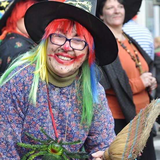 Eine Person verkleidet als Hexe mit mehrfarbigem Haar, trägt einen schwarzen Hut, eine Brille und hält einen Besen. Sie lächelt und scheint sich auf einem Halloween-Event zu befinden.