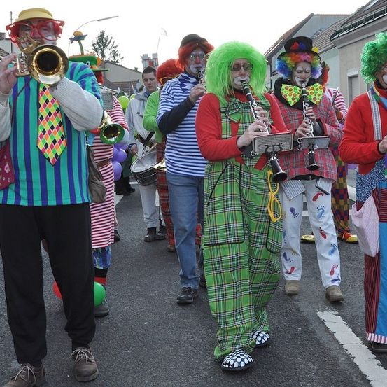 Eine Gruppe von Clowns in bunten Kostümen spielt Musikinstrumente bei einer Parade. Sie halten Trompeten, Klarinetten und Trommeln.