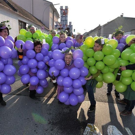Eine Gruppe von Menschen in Traubenkostümen mit lila und grünen Luftballons, die für ein Foto auf einer Straße posieren.