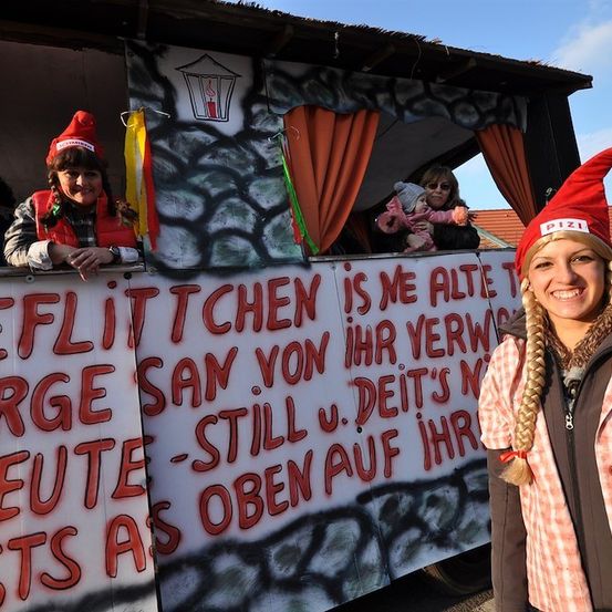 Eine Frau mit rotem Hut steht neben einem geschmückten Wagen mit einem Banner, auf dem 'Fleischlichen is ne alte T' und andere Texte stehen. Eine weitere Frau im Wagen hält ein Baby.