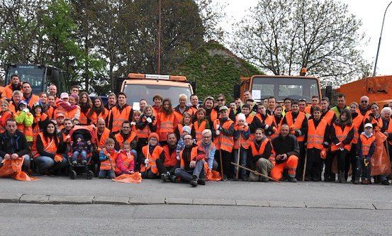 Eine große Gruppe von Menschen in orangenen Westen und Handschuhen posiert für ein Foto auf einer Straße mit orangefarbenen Maschinenfahrzeugen im Hintergrund.