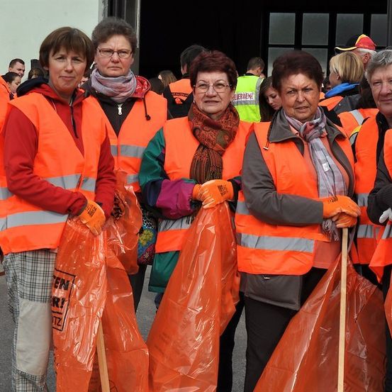 Eine Gruppe von Frauen in orangenen Westen, die orange Müllsäcke und Stöcke halten, steht vor einem Gebäude.