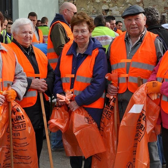 Eine Gruppe älterer Freiwilliger in orangefarbenen Westen, die orangefarbene Taschen und Stöcke halten, möglicherweise für eine Gemeinschaftsreinigungsveranstaltung.