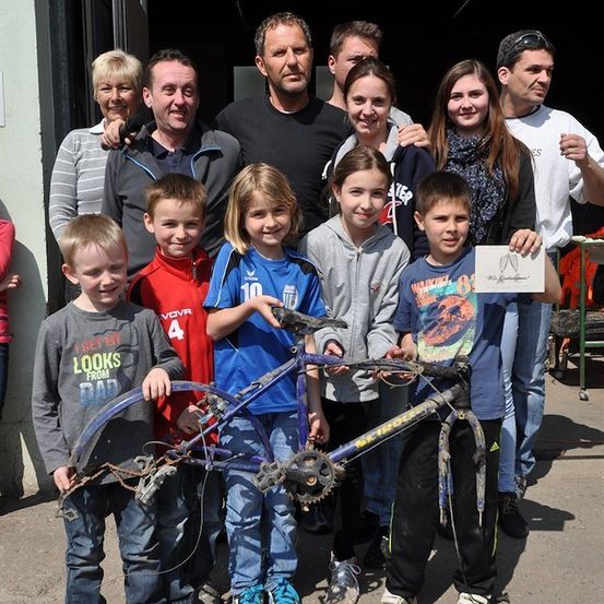 Eine Gruppe von Menschen, einschließlich Kindern, posiert für ein Foto mit einem beschädigten Fahrrad vor einem Gebäude.