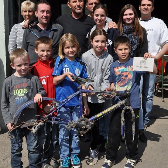 Eine Gruppe von Menschen, einschließlich Kindern, steht draußen und hält ein zerlegtes Fahrrad. Sie lächeln und posieren für ein Foto.