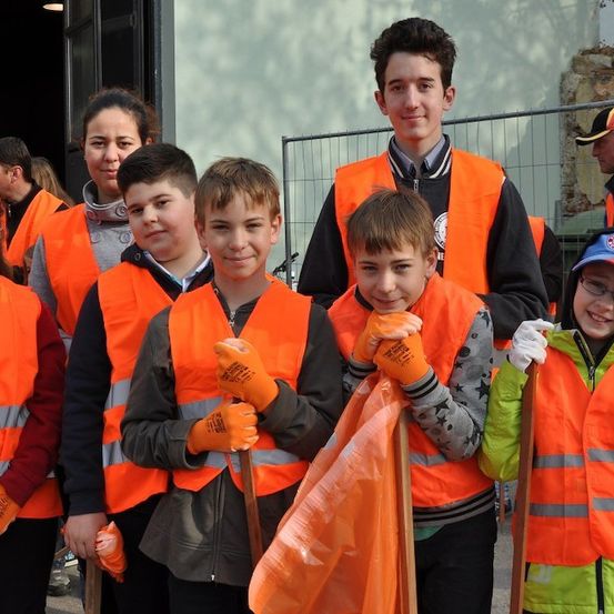 Eine Gruppe von Kindern und Erwachsenen in orangefarbenen Sicherheitswesten, die Stöcke und Taschen halten, wahrscheinlich an einer Gemeinschaftsreinigungsveranstaltung teilnehmend.