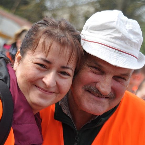 Ein Mann und eine Frau lächeln und posieren für ein Foto. Sie tragen beide orange Westen. Der Mann trägt einen weißen Hut. Hinter ihnen sind verschwommene Menschen und Bäume.