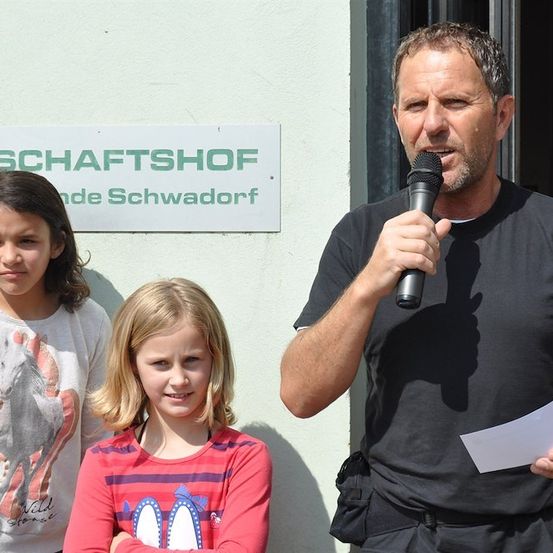 Ein Mann spricht in ein Mikrofon und hält ein Papier in der Hand, neben zwei jungen Mädchen vor einem Schild.