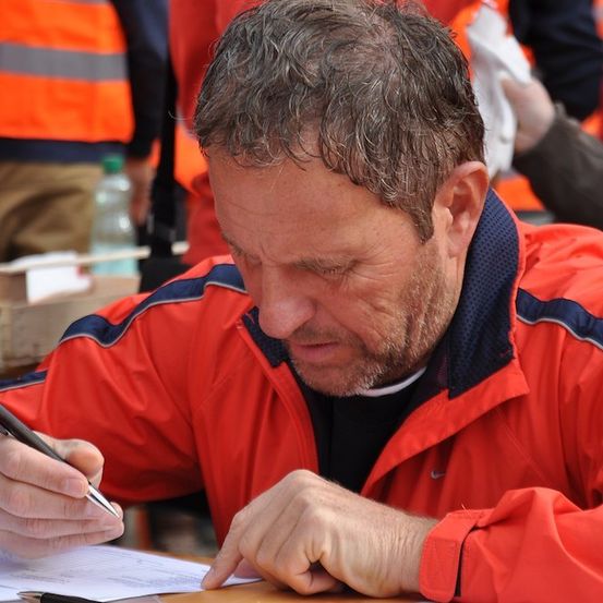 Ein Mann in einer orangefarbenen Jacke unterzeichnet ein Dokument und schaut nach unten, mit verschwommenen Personen in orangefarbenen Westen im Hintergrund.