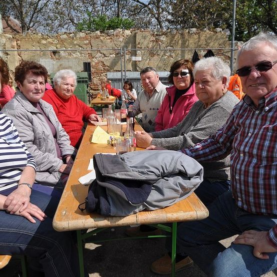 Eine Gruppe älterer Menschen sitzt an einem Tisch in einem Außenbereich und posiert für ein Foto. Sie tragen Freizeitkleidung und einige von ihnen tragen eine Brille. Auf dem Tisch befinden sich Gläser, Flaschen und eine Tasche. Hinter ihnen steht eine Steinmauer mit einem Metallzaun.