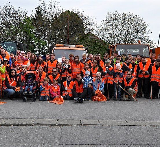 Eine große Gruppe von Menschen in orangefarbenen Westen und Handschuhen posiert für ein Foto vor einer Straße mit Maschinenfahrzeugen im Hintergrund.