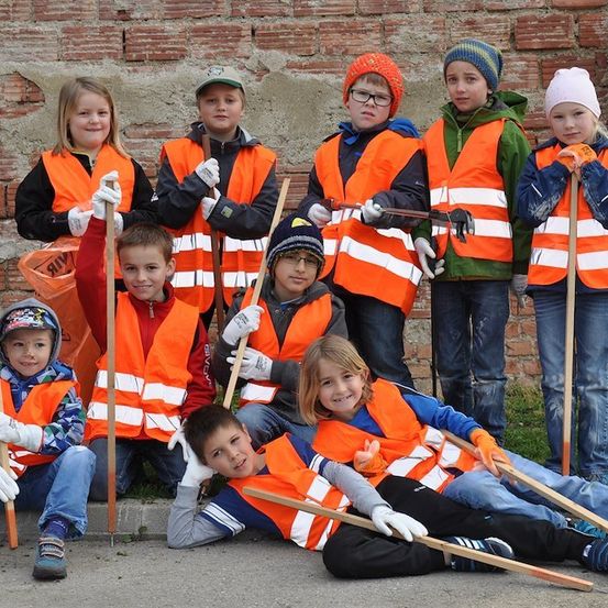 Eine Gruppe von Kindern in orangefarbenen Westen und Handschuhen steht und sitzt auf dem Boden, hält Stöcke und posiert für ein Foto vor einer Ziegelmauer.