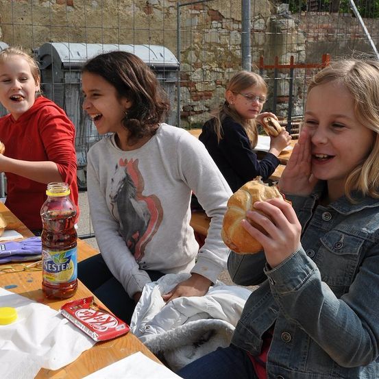 Vier Mädchen sitzen an einem Tisch und essen Sandwiches und trinken. Ein Mädchen hält ein Sandwich und lacht. Ein anderes Mädchen hält ein Sandwich und trinkt aus einem Becher. Eine Flasche Nestlea steht auf dem Tisch.