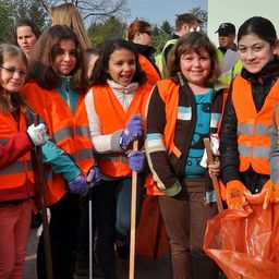 Eine Gruppe junger Mädchen in orangefarbenen Westen und Handschuhen lächelt und posiert für ein Foto, während sie Stöcke und einen Plastikbeutel halten.