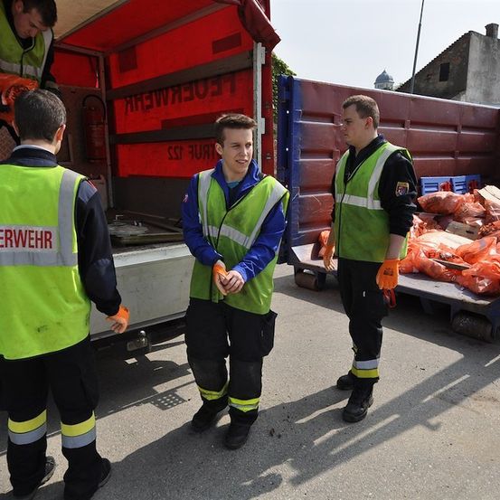 Drei Feuerwehrleute in grünen Westen stehen vor einem roten LKW, einer hält einen Feuerlöscher. Der LKW ist mit orangefarbenen Säcken beladen.