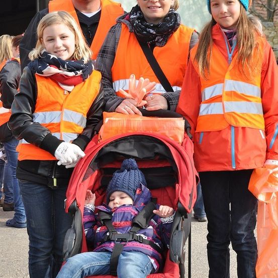 Eine Familie mit vier Personen, darunter zwei junge Mädchen und ein Baby im Kinderwagen, gekleidet in orangefarbenen Westen, stehen zusammen draußen.