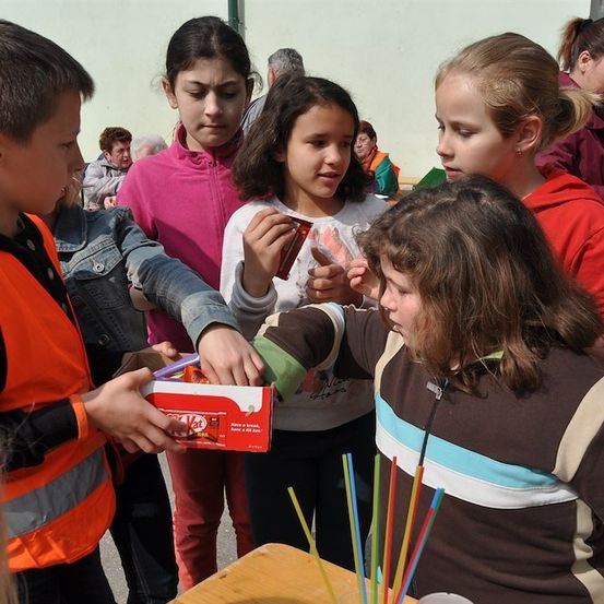 Eine Gruppe von Kindern ist um einen Tisch versammelt, wobei ein Kind eine Schachtel mit Süßigkeiten hält und ein anderes Kind danach greift. Die Kinder tragen Jacken und einige haben Pferdeschwänze. Im Hintergrund stehen andere Menschen und schauen die Kinder an.