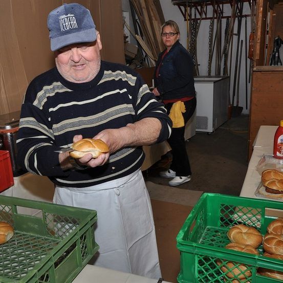 Ein Mann in einem gestreiften Pullover und einer blauen Kappe hält ein Sandwich in einer Küche, während eine Frau hinter ihm steht. Es gibt Tabletts mit Sandwiches, eine Ketchupflasche und verschiedene Küchenutensilien im Hintergrund.