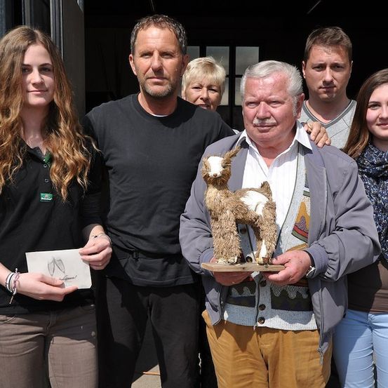 Eine Gruppe von Menschen steht zusammen und lächelt für ein Foto. Ein Mann hält eine Trophäe eines Ziegenbocks, während eine Frau ein Stück Papier in der Hand hält. Dahinter stehen eine weitere Frau und ein Mann dicht beieinander.