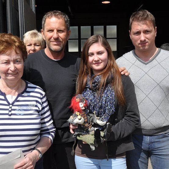 Eine vierköpfige Familie steht vor einem Gebäude, wobei eine Frau eine Puppe hält und ein Mann ein Stück Papier in der Hand hat.