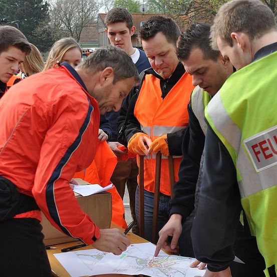 Eine Gruppe von Menschen in orangefarbenen Westen versammelt sich um einen Tisch mit einer Karte, möglicherweise um einen Plan zu besprechen. Ein Mann zeigt auf die Karte, während andere sie betrachten.