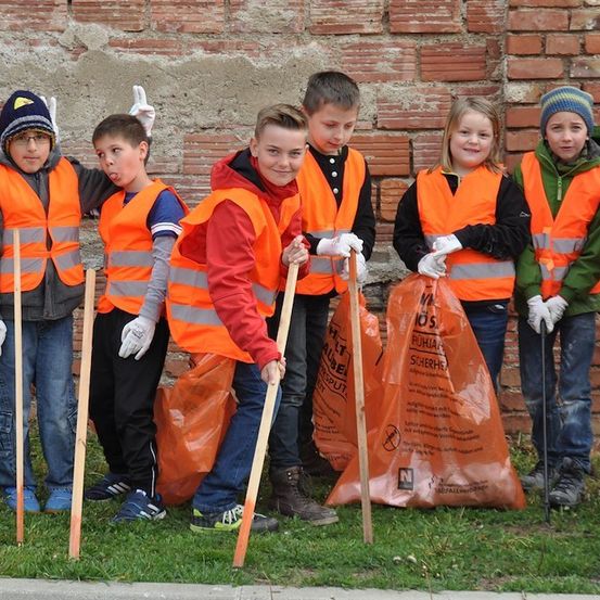 Eine Gruppe von Kindern in orangefarbenen Westen und Handschuhen steht auf einer Rasenfläche und hält Stöcke und orangefarbene Taschen. Sie scheinen an einer Gemeinschaftsaktion zur Müllentsorgung teilzunehmen.
