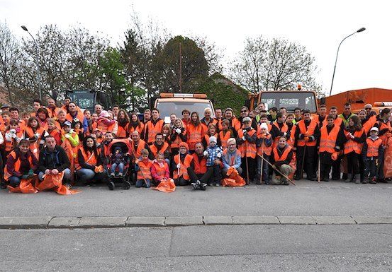 Eine große Gruppe von Menschen in orangefarbenen Westen und Handschuhen posiert für ein Foto auf einer Straße, mit Maschinenfahrzeugen und Bäumen im Hintergrund.