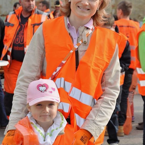 Eine Frau in einer orangefarbenen Warnweste steht neben einem kleinen Mädchen in einer rosa Mütze und orangefarbenen Handschuhen und lächelt in die Kamera. Dahinter stehen mehrere Personen in orangefarbenen Warnwesten.