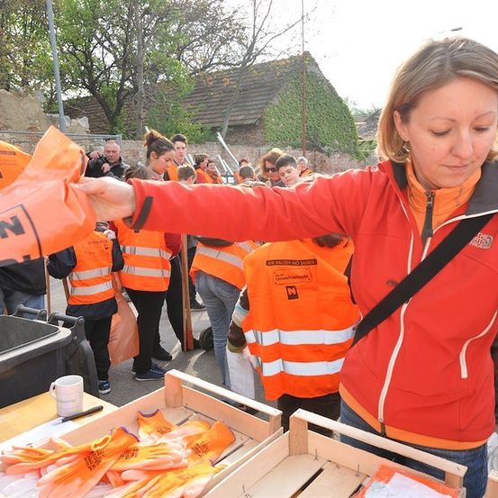 Eine Frau in einer orangefarbenen Weste verteilt orange Handschuhe an eine Gruppe von Menschen in passenden Westen. Sie stehen vor einem Tisch mit Handschuhen und einer Tasse. Bäume und Gebäude sind im Hintergrund.