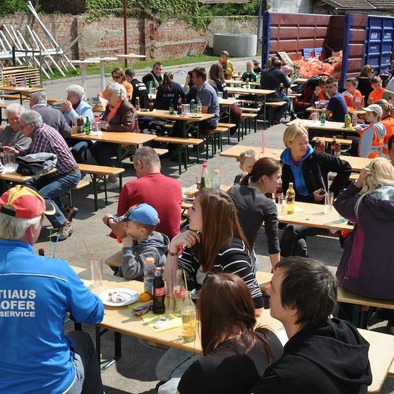 Eine Gruppe von Menschen sitzt an Tischen in einem Außenbereich, möglicherweise bei einem Festival. Einige tragen Hüte und Brillen, und einige halten Flaschen. In der Ferne gibt es einen blauen Müllcontainer und eine Ziegelmauer.