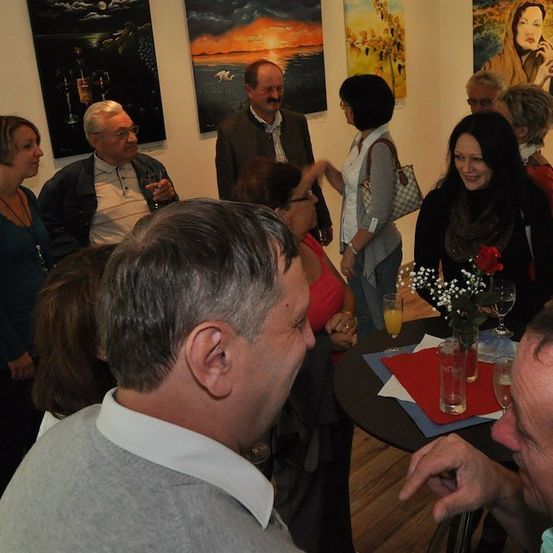 Eine Gruppe von Menschen versammelt sich in einer Kunstgalerie und unterhält sich. Im Vordergrund steht ein Tisch mit einem Blumenstrauß und Weingläsern.