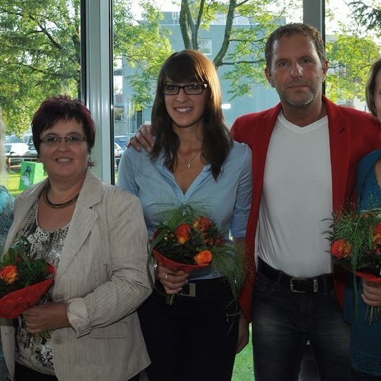 Fünf Erwachsene, drei Frauen und zwei Männer, stehen zusammen und halten Blumen. Sie lächeln und scheinen für ein Foto zu posieren.
