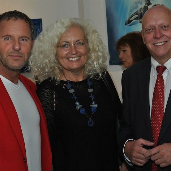 Drei Menschen stehen zusammen in einem Raum und lächeln für ein Foto. Der Mann auf der linken Seite trägt einen roten Mantel, die Frau in der Mitte hat weiße Haare und trägt eine Brille, und der Mann auf der rechten Seite trägt einen Anzug und eine Krawatte. Hinter ihnen ist eine Wand mit einem Wal-Gemälde.