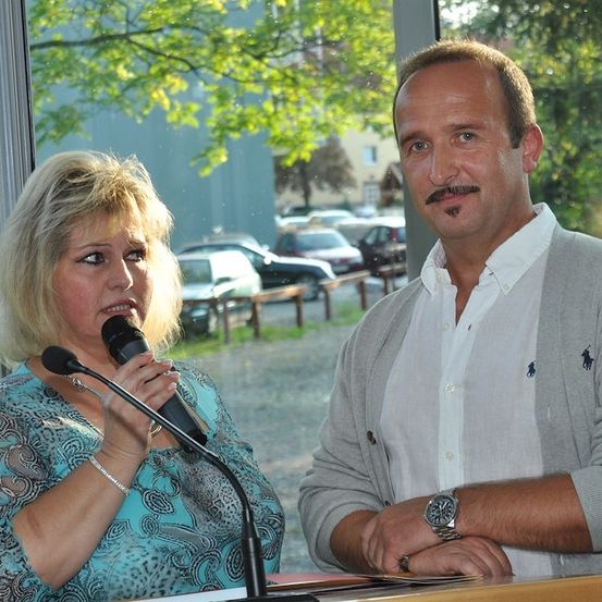 Eine blonde Frau spricht in ein Mikrofon an einem Rednerpult, während ein Mann mit Schnurrbart neben ihr steht und beide in die Kamera schauen. Hinter ihnen zeigt ein Fenster einen Parkplatz mit Autos und Bäumen.