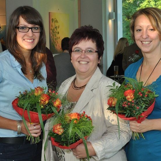 Drei Frauen in einem Raum, zwei halten Blumensträuße und lächeln für ein Foto. Die Frau in der Mitte trägt eine Brille und eine Halskette.