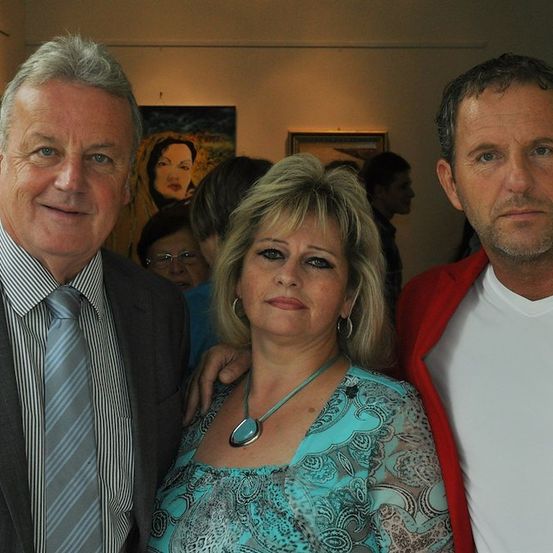 Drei Menschen stehen in einer Kunstgalerie zusammen, wobei zwei Männer eine Frau flankieren. Der Mann links trägt einen gestreiften Anzug und eine Krawatte, während der Mann rechts eine rote Jacke trägt. Die Frau trägt ein gemustertes Oberteil und eine Halskette.
