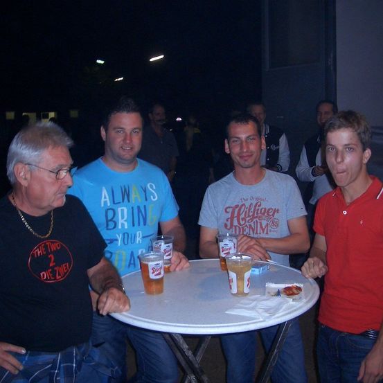 Vier Männer stehen um einen Tisch mit Getränken, zwei von ihnen tragen Brillen und einer raucht. Auf dem Tisch stehen Biergläser und eine Schachtel Zigaretten.