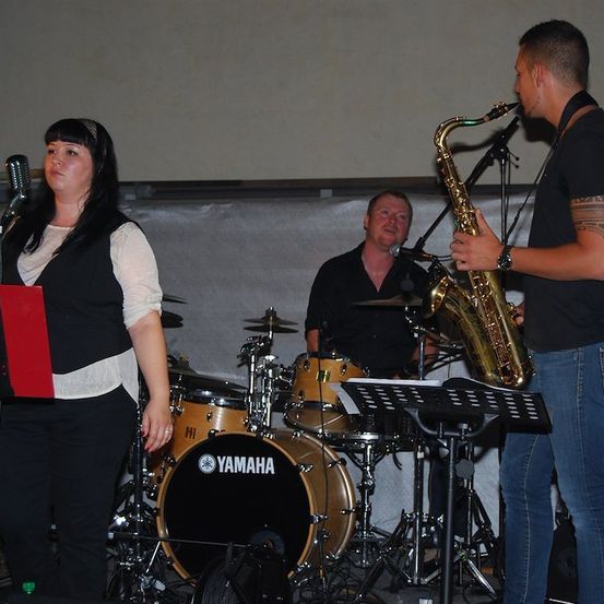 Eine Frau singt in ein Mikrofon, während ein Mann Saxophon spielt und ein anderer Mann Schlagzeug spielt.