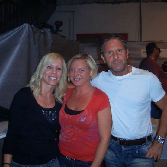 Drei Personen, zwei Frauen und ein Mann, stehen zusammen und lächeln für ein Foto. Die Frau links trägt ein schwarzes Oberteil, die mittlere Frau trägt ein rotes Oberteil und der Mann trägt ein weißes Hemd und eine Uhr.