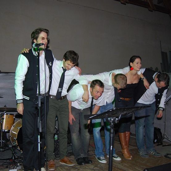 Eine Gruppe von Menschen auf der Bühne mit Musikinstrumenten, ein Mann singt in ein Mikrofon, während andere sich an ihn lehnen, und ein Schlagzeug im Hintergrund.