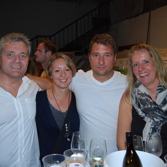 Vier Menschen lächeln und posieren für ein Foto bei einer gesellschaftlichen Zusammenkunft. Der Mann auf der linken Seite trägt ein weißes Hemd, während die Frau auf der rechten Seite ein schwarzes Top und einen Schal trägt. Der Mann in der Mitte trägt ein weißes T-Shirt. Hinter ihnen stehen Weingläser und eine Flasche Wein.