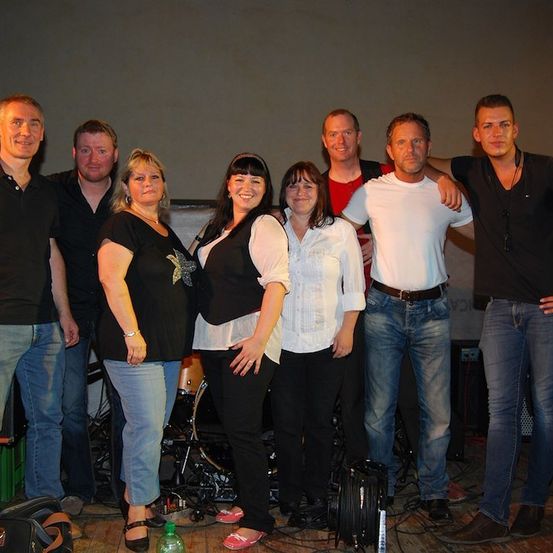 Eine Gruppe von Musikern, sowohl Männer als auch Frauen, stehen zusammen in einem Studio mit Musikinstrumenten im Hintergrund.